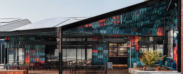 Exterior view of a contemporary commercial hospitality venue with a dramatic sloping roof and extensive black metal framing. The facade is clad in a vivid vertical multicolour pattern of teal, blue, red and grey panels, with large glazed openings revealing dining and bar areas inside. Outdoor tables and chairs occupy the forecourt, while a planted raised area and seating appear near the entrance.