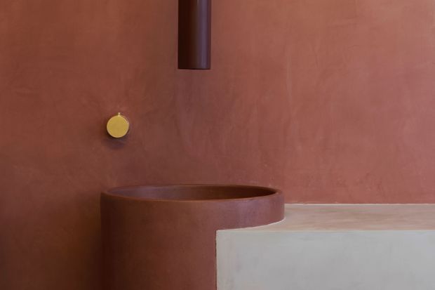 Close-up architectural detail of a minimalist bathroom washbasin against a terracotta-toned plaster wall. A cylindrical dark metal tap rises above a rounded integrated basin, with a small circular brass-gold control. A pale stone or concrete vanity surface occupies the foreground.