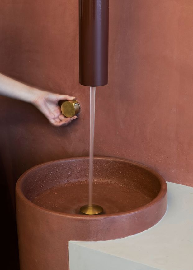 Close-up of a sculptural bathroom basin in a warm terracotta finish, with a matching cylindrical wall-mounted faucet and brass-toned controls and drain. The round vessel sink is integrated into a curved pedestal-like surround and sits beside a pale solid-surface or stone counter. Water is running, emphasizing the tactile, monolithic design.