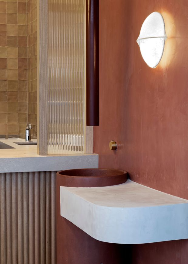 A close-up view of a highly designed bathroom featuring a sculptural white vessel basin on a rounded projecting vanity, a warm terracotta-toned wall, a ribbed vertical timber or plaster finish below the adjacent counter, and a translucent fluted glass screen. A wall-mounted sculptural light and brass-toned tapware add refined decorative details. The background includes a second basin and tiled wall.