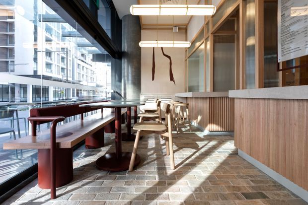 A bright, contemporary commercial café or dining space with floor-to-ceiling glazing, built-in timber banquette seating, timber tables and stools, a long wood-clad service counter, and exposed concrete wall surfaces. Linear pendant lights, pale timber screens, and patterned masonry flooring create a warm, refined interior. Urban apartment buildings are visible through the glazing.