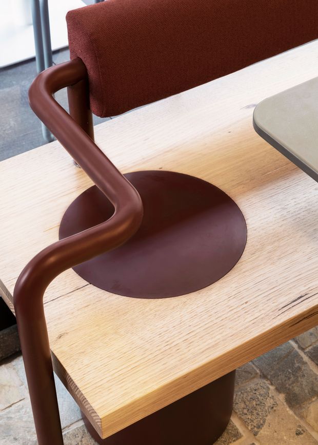 Close-up product-style image of a modern chair beside a light timber table. The chair features a curved dark brown tubular frame and circular seat base, with a rust-red upholstered backrest. A pale rounded tabletop or adjoining surface is visible at the upper right. The image focuses on furniture materials, joinery, and warm neutral colours.
