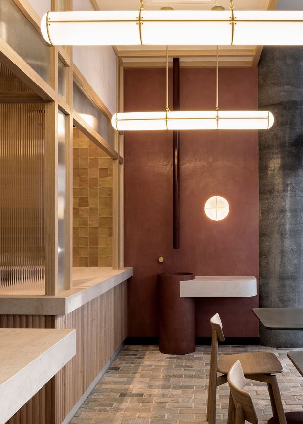 A refined hospitality-style interior featuring a compact bar or dining area with warm timber screening and cabinetry, textured translucent glazing, a deep terracotta plaster feature wall, dark stone or plaster surfaces, and suspended elongated linear pendant lights with brass detailing. A sculptural round basin and floating pale stone counter sit against the feature wall, while a small dining table and timber chairs occupy the foreground. The floor is finished in small rectangular brick-like tiles, creating a warm, tactile and richly layered atmosphere.