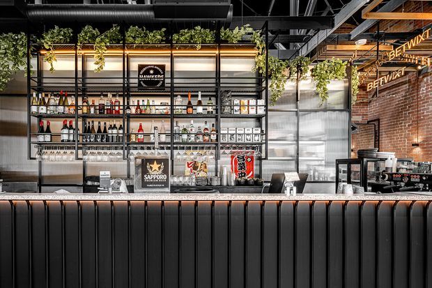 A commercial bar counter with a Japanese-inspired drinks and service area. The space features black vertically fluted counter panelling, stainless-steel work surfaces and shelving, exposed black-painted ceiling services, hanging greenery, warm linear lighting, and a partially exposed red-brick wall. Bottles, glassware, coffee equipment, and Japanese beverage signage are arranged behind the counter.