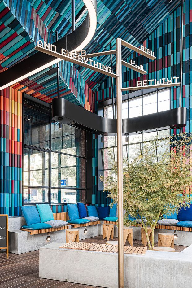 A vibrant contemporary commercial interior with a tall glazed facade, dramatic layered geometric ceiling and wall cladding in teal, turquoise, red, pink and dark blue. A sculptural black suspended light wraps around a central freestanding signpost with directional text. Built-in concrete seating platforms with bright blue cushions surround low timber-topped concrete tables, while an indoor tree adds greenery. The space has an energetic, creative hospitality or workplace-lounge character.