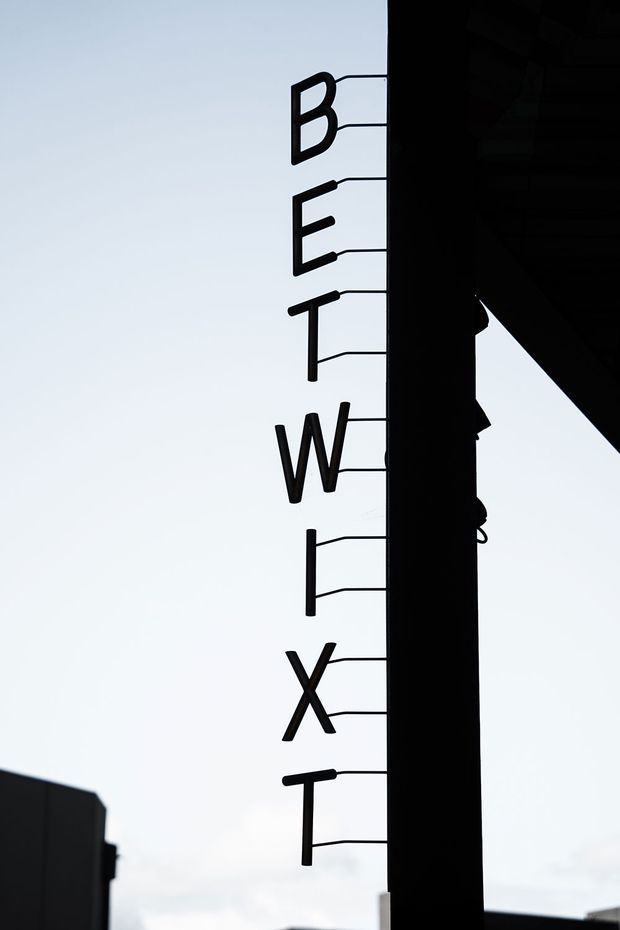 A stark, vertically oriented exterior architectural detail showing black steel lettering and thin horizontal linework mounted beside a tall dark structural column or facade edge. The letters appear to form part of a building sign, silhouetted against a pale sky, with angular roof or canopy elements visible at the right and lower edges. The image has a minimalist, high-contrast, graphic quality.