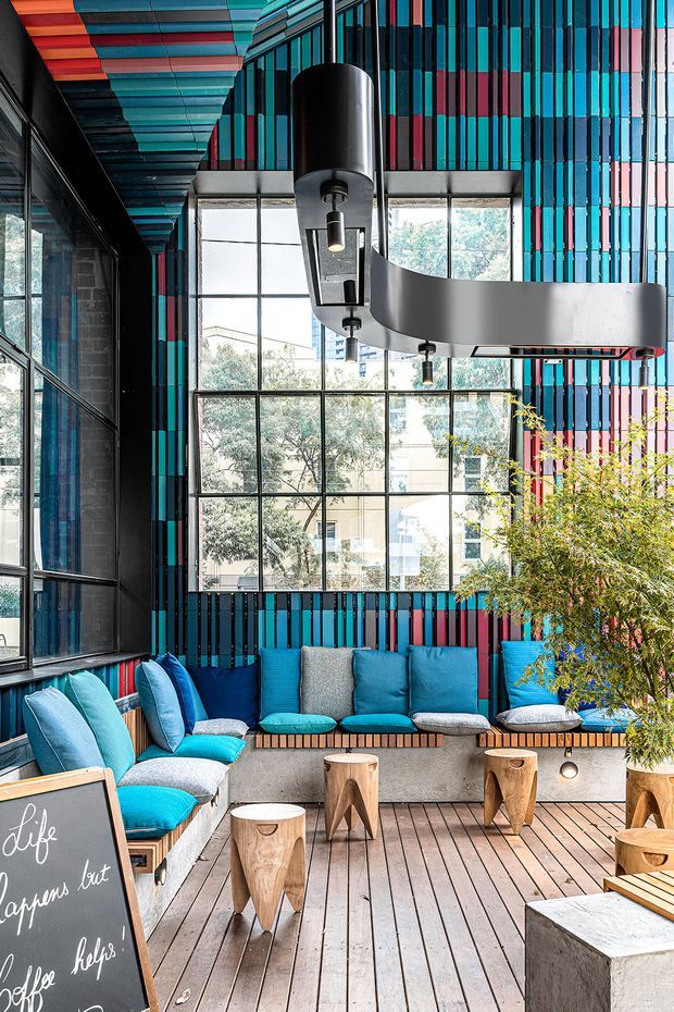 A vibrant contemporary indoor lounge or café-style breakout area with extensive black-framed glazing, a colourful striped wall and ceiling installation in turquoise, blue, pink and red, built-in grey upholstered bench seating with bright blue cushions, small timber stools and tables, and a large sculptural suspended light fitting. A planted tree adds greenery beside the seating.