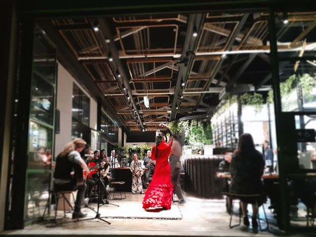 A lively indoor hospitality venue, likely a restaurant or café, viewed through a dark-framed glazed entrance. Diners sit at tables along both sides while a performer in a vivid red dress stands in the central aisle. The space features a dramatic exposed services and lighting ceiling with dark structural framing, linear timber-toned slats, metal conduits, and large glazed openings. Greenery is visible beyond the glazing, contributing to an indoor–outdoor atmosphere.