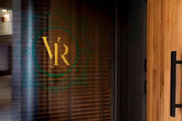 Close-up of a refined entry door featuring warm vertical timber cladding, a dark metal frame and handle, and a glass panel with timber venetian blinds. The glass bears a prominent gold monogram logo with a decorative green circular motif, suggesting a branded hospitality or commercial entrance.