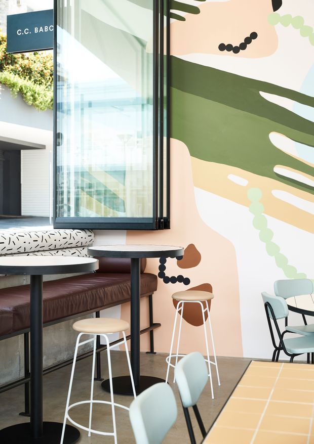 A brightly lit contemporary commercial café or hospitality interior with floor-to-ceiling glazing, built-in leather banquette seating, round café tables, stools, and loose dining chairs. A large abstract mural in soft peach, sage green, cream, and black dominates the wall, while a hanging sign is visible near the glazed entry.