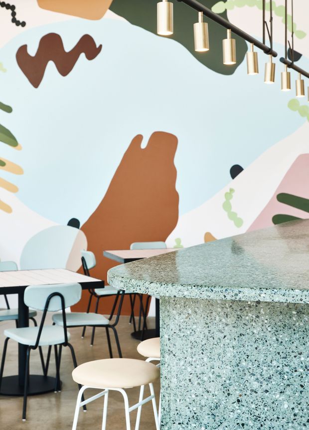 A playful contemporary café interior featuring a large colourful abstract mural, pale blue and white furniture with black metal frames, and a prominent curved terrazzo counter in the foreground. A linear arrangement of cylindrical pendant lights hangs from a black ceiling track, while small dining tables and stools create a casual hospitality setting.