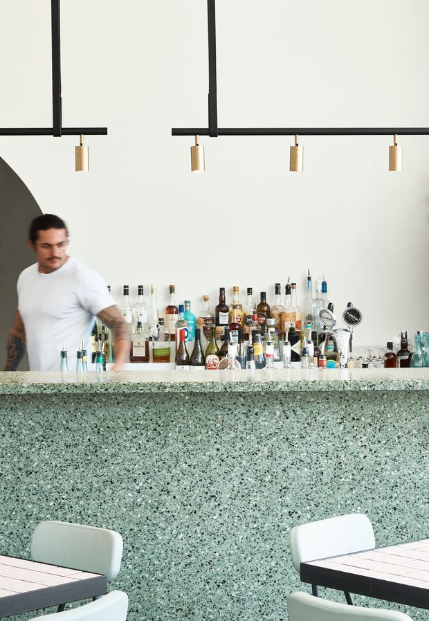 A contemporary bar interior with a long terrazzo-faced counter and matching pale speckled benchtop. Bottles and bar equipment line the rear counter, with a bartender behind it. Black linear pendant lighting with small brass-toned drop lights hangs overhead, while dining tables and pale chairs occupy the foreground. The space has a clean, graphic, modern hospitality aesthetic.