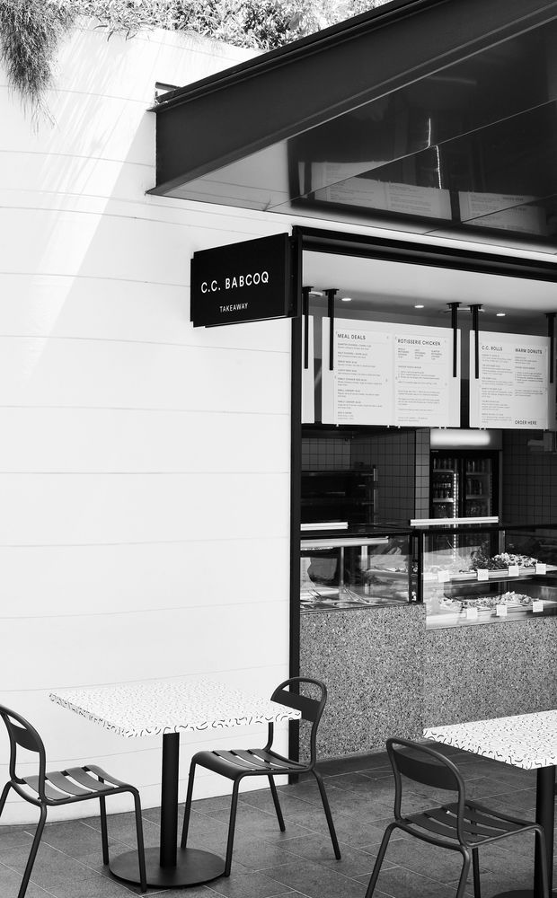 Black-and-white photograph of a compact takeaway barbecue restaurant storefront, with a projecting C.C. Babcoq sign, open service counter, menu boards, tiled interior, display counter and outdoor café seating on a paved terrace. The facade combines white horizontal cladding with dark metal framing and a polished terrazzo-like counter front.
