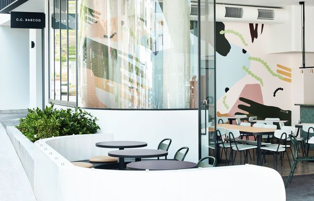 Bright contemporary commercial café interior with a semi-enclosed outdoor-style seating area, round tables, green metal chairs, built-in white planter seating, extensive glass partitions, and a large abstract mural. The space has polished concrete flooring, white surfaces, black accents, and abundant natural light.