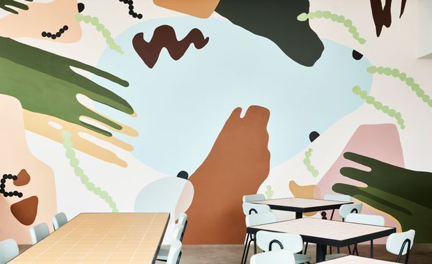 A brightly coloured contemporary dining or café interior with large abstract mural artwork covering the walls. Several light tables with black edging and white chairs fill the space, with a warm tan tiled table in the foreground and a neutral hard floor. The mural uses organic shapes in pale blue, cream, green, brown, pink and black, creating a playful, graphic atmosphere.