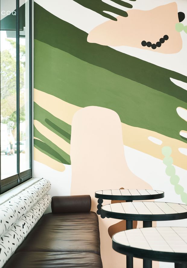 A bright contemporary interior corner with floor-to-ceiling glazing, a dark brown leather banquette, round nesting tables with black metal frames and pale tiled tops, and a large abstract mural in olive green, cream, peach and black. The space has a playful, graphic character and abundant natural light.