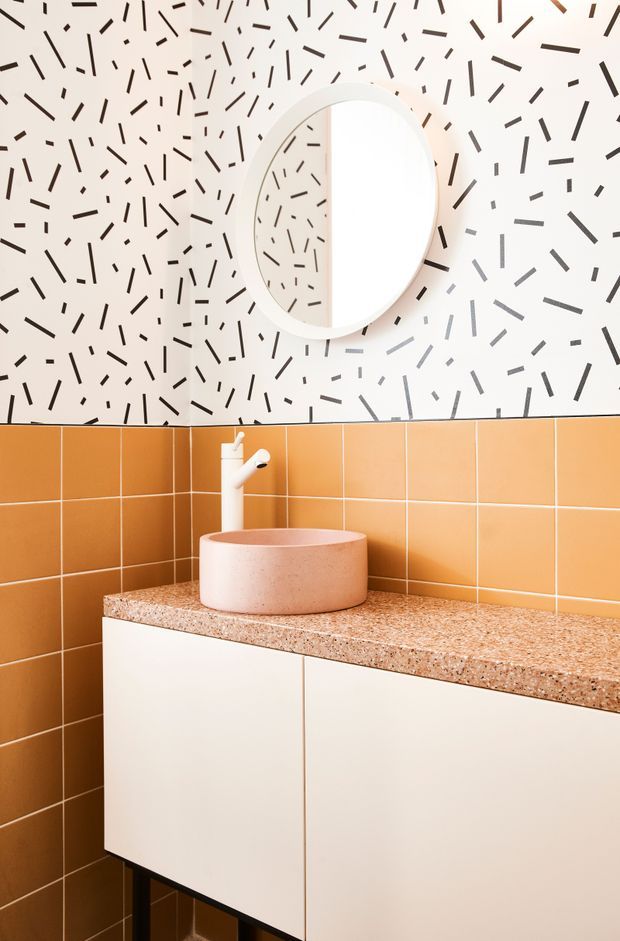 A compact, brightly styled bathroom vanity featuring a round pink vessel basin, white wall-mounted faucet, white floating cabinetry, and a terrazzo benchtop. The lower walls are clad in warm orange square tiles, while the upper wall has a playful white-and-black linear patterned finish. A large round mirror is mounted above the basin.
