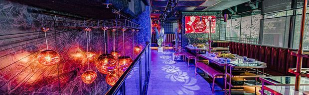 Vividly lit commercial dining or bar interior with a long banquette along the right wall, individual tables and chairs, dramatic sculptural amber pendant lights, patterned wall graphics, reflective surfaces, and saturated purple, blue, and red lighting. The space has an eclectic, theatrical atmosphere and appears designed for hospitality use.