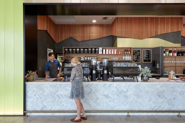 A modern café or hospitality counter viewed from the customer area. The long front counter has a pale grey herringbone-patterned finish, with a dark countertop and brass-toned foot rail. Behind it are coffee machines, grinders, display shelving, drinks, and menu boards. The backdrop combines dark textured geometric wall panels with warm vertical timber slats, framed by a bright lime-green wall and black overhead surround. The space is brightly lit and contemporary, with staff and a customer at the counter.