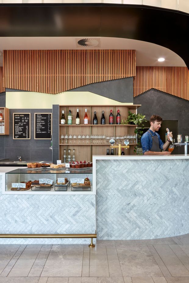 Interior photograph of a contemporary café or hospitality counter. The space features a pale stone or marble-patterned herringbone counter front, dark grey tiled wall surfaces, warm timber shelving and vertical timber slat detailing, integrated bottle display, glassware, taps, and a black curved ceiling bulkhead. A staff member is serving behind the counter.