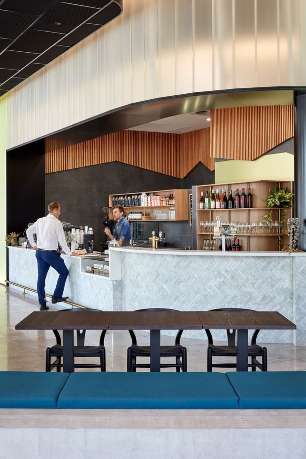 A contemporary commercial hospitality interior featuring a bar and café service counter beneath a dramatic curved black ceiling canopy. The bar front is clad in pale herringbone-patterned stone or tile, with dark textured wall surfaces, warm vertical timber slat detailing, open timber bottle shelving, and integrated lighting. A communal dark timber table with black stools and a long blue upholstered bench occupy the foreground.