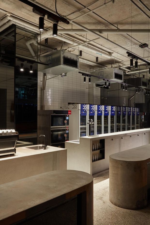A dimly lit contemporary commercial hospitality interior, likely a café, bar, or food-service venue. The space features exposed concrete ceilings with visible services and ductwork, suspended black track lighting, white tiled walls, stainless-steel ventilation units, commercial ovens, refrigerated display cabinets, and substantial pale concrete counters and cylindrical tables. The fit-out has an industrial character with dark reflective glazing and strong blue accents on the refrigerated units.