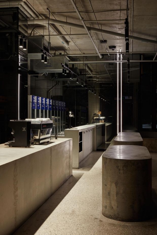A dark, industrial-style café or commercial hospitality interior with exposed concrete ceilings, visible services and ductwork, polished concrete counters, cylindrical concrete tables, black metal detailing, and dramatic linear pendant lighting. A coffee machine and service counter are visible in the foreground, with reflective glazing and additional seating extending into the space.