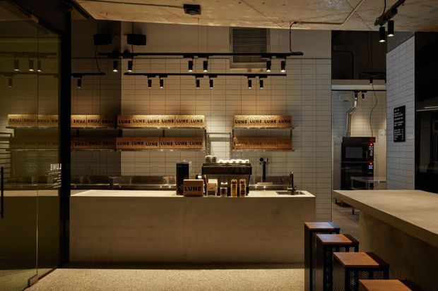 A dimly lit contemporary commercial café or food-service interior with a long pale stone or concrete service counter, tiled walls, open shelving stocked with branded supplies, espresso equipment, stools, and exposed ceiling services. Track lighting and black-framed glazing create an industrial, refined atmosphere.