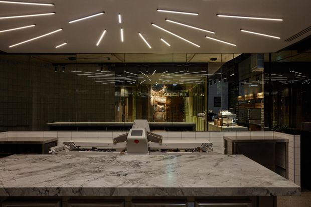 A dark, refined commercial interior viewed from behind a large veined stone service counter or bar. The foreground features a broad marble or natural-stone benchtop with integrated commercial equipment, while the rear wall is lined with reflective glazing and dark tiled or stone surfaces. Linear recessed ceiling lights radiate across the ceiling and are repeated in reflections, creating a dramatic geometric lighting effect. Shelving, counters, and equipment are visible beyond the glazed partition.