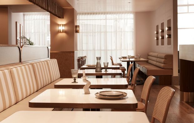 Warm, refined restaurant or café interior with rows of cream-and-tan upholstered banquette seating, white tabletops with dark wood edges, woven dining chairs, decorative wall lighting, fluted timber detailing, and large sheer-covered windows providing soft daylight. The space has a retro-inspired, polished hospitality fit-out with a long communal dining arrangement.