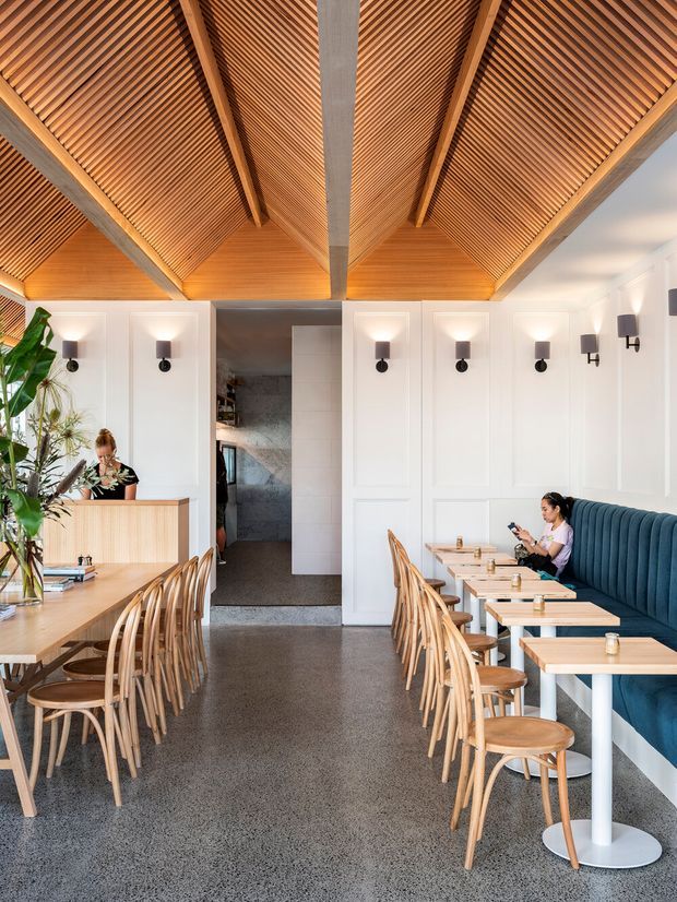 Bright contemporary café or restaurant interior with rows of light timber dining tables and curved wooden chairs, a long deep-blue upholstered banquette, white decorative wall panelling, and evenly spaced black wall sconces. The space features a dramatic vaulted ceiling lined with warm timber slats and exposed timber beams, with polished grey concrete or terrazzo flooring. A service counter and staff member are visible at the rear-left, while a central opening leads to a darker back-of-house area.