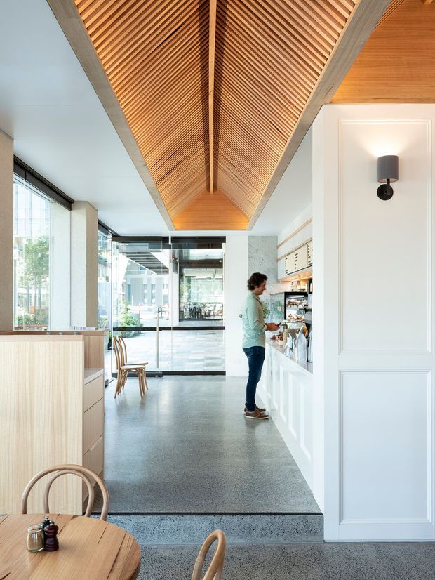 Bright contemporary café interior with a long white service counter and integrated shelving, pale timber cabinetry, expansive glass doors and windows, and a polished grey concrete or terrazzo floor. The main architectural feature is a dramatic pitched ceiling lined with warm timber slats and exposed grey structural beams. A small dining area occupies the foreground, while the glazed frontage opens onto an urban setting.