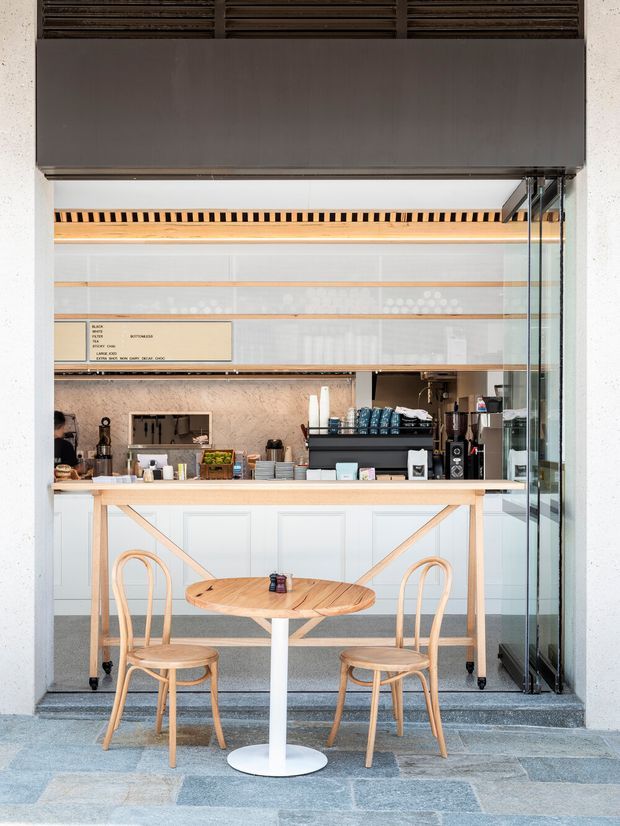 A compact contemporary café or takeaway service space viewed from the entrance, with a white and pale timber counter, espresso machine, menu board, shelves, and a small round café table with bentwood chairs in the foreground. The storefront is framed by glass doors and a dark overhead awning or bulkhead, with slate-like stone paving at the threshold.