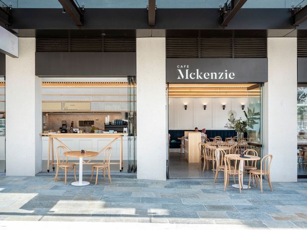 Street-facing view of Cafe McKenzie, a contemporary café with an open glazed frontage. The façade features broad white structural columns, dark louvred ventilation panels, a dark projecting canopy with exposed beams, and large glass openings. Inside are a service counter, light timber café tables and chairs, pendant-style wall lights, banquette seating, and greenery. The forecourt is finished with large-format grey stone paving.