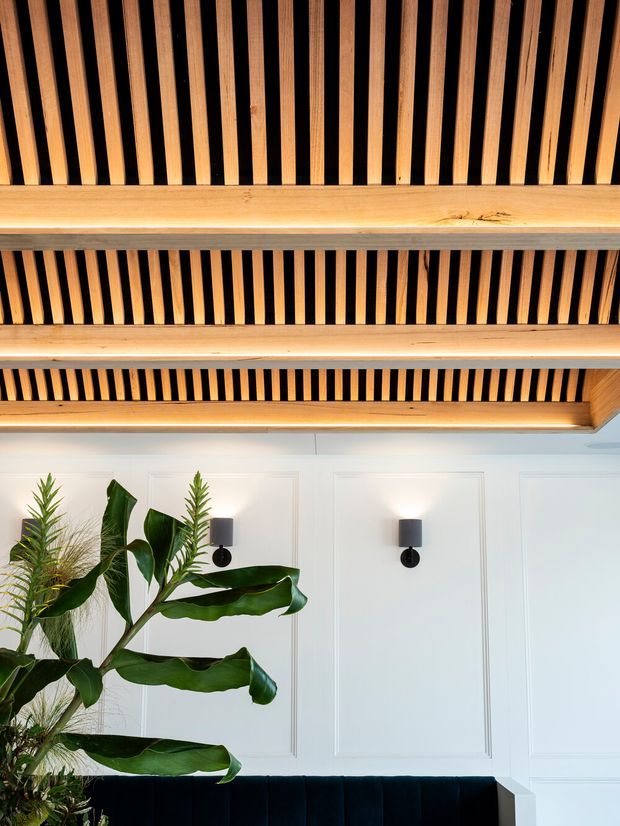 Interior detail showing a white wall with decorative rectangular panel moulding, paired black wall sconces, and the upper edge of a dark upholstered sofa. A striking ceiling features exposed warm timber battens arranged in parallel rows with dark recessed gaps, integrated linear lighting, and substantial timber beams. Tropical foliage occupies the lower left foreground. The composition has a refined contemporary character with strong timber detailing and contrasting black accents.