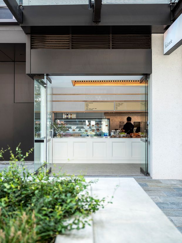 Street-facing storefront of a small modern café or food-service counter, viewed from an outdoor walkway. The open glazed frontage reveals a white paneled service counter, refrigerated display case, menu boards, shelving, and staff inside. Dark metal framing and a projecting canopy create a contemporary commercial facade, with low landscaping in the foreground.