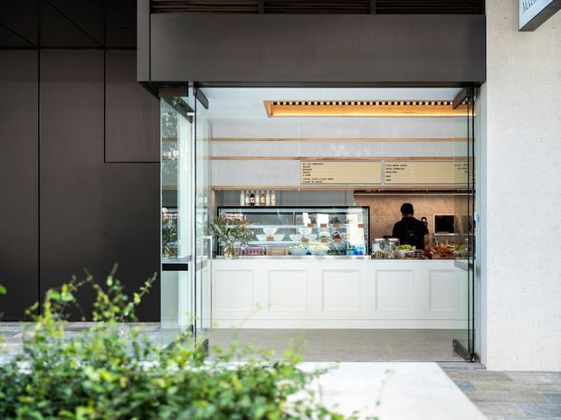 A street-facing commercial café or food-service counter viewed through an open glass frontage. The space features a white paneled service counter, glass display case with prepared food, timber-toned shelving and menu boards, dark exterior cladding, and a contemporary recessed ceiling light. Landscaping is visible in the foreground.
