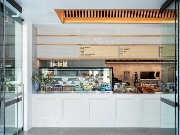 A bright, compact contemporary café or takeaway counter viewed from the entrance. The space features a white paneled service counter with glass food displays, a warm timber-toned tiled or textured backsplash, pale timber menu boards, and a striking recessed ceiling feature with timber slats and integrated lighting. Glass entry doors frame the scene on both sides.