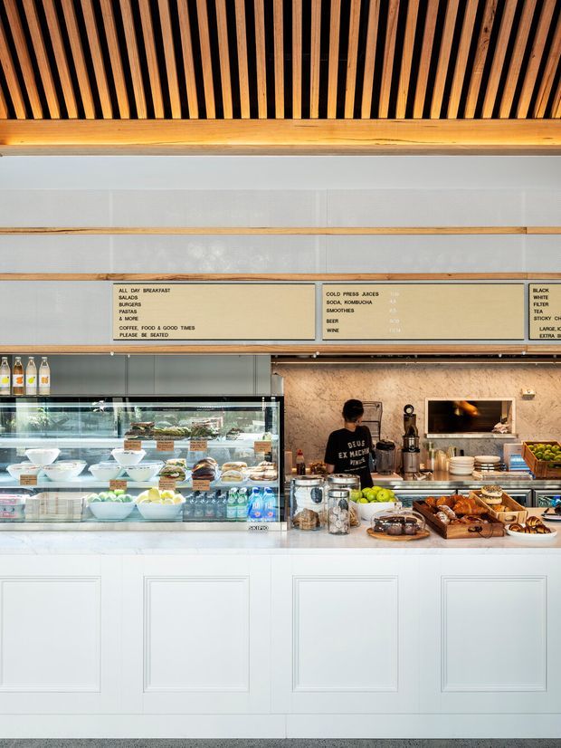 A bright contemporary café counter interior with a long white paneled service counter, glass refrigerated display filled with pastries, desserts and drinks, timber shelving and signage, and a warm slatted timber ceiling with exposed beams. Behind the counter are food-preparation areas, appliances and shelving, creating a clean yet warm hospitality setting.