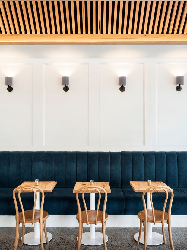 A refined hospitality-style dining area with a long deep navy upholstered banquette beneath evenly spaced wall sconces. Three small light timber tables with curved timber café chairs and white pedestal bases line the seating. The white walls feature decorative rectangular panel moulding, while a slatted timber ceiling feature adds warmth and texture. The space has a clean, symmetrical, contemporary-classic appearance.
