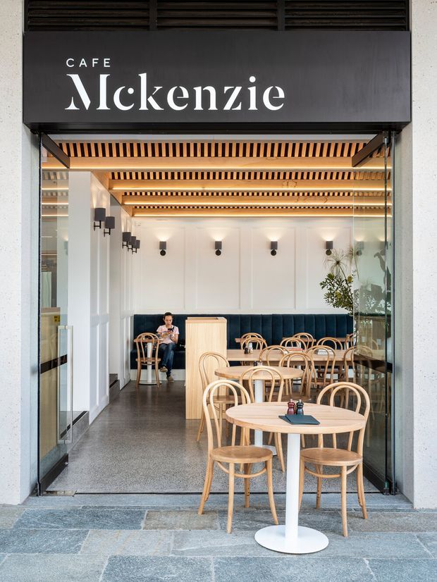 Front-facing view into Cafe McKenzie, a compact contemporary cafe with a dark charcoal sign and full-height glazed entrance. The interior features white paneled walls, a warm timber slatted ceiling with integrated lighting, dark terrazzo-like flooring, built-in dark upholstered banquette seating, and light timber café tables and bentwood chairs. Outdoor seating is positioned directly outside the entrance on grey stone paving.