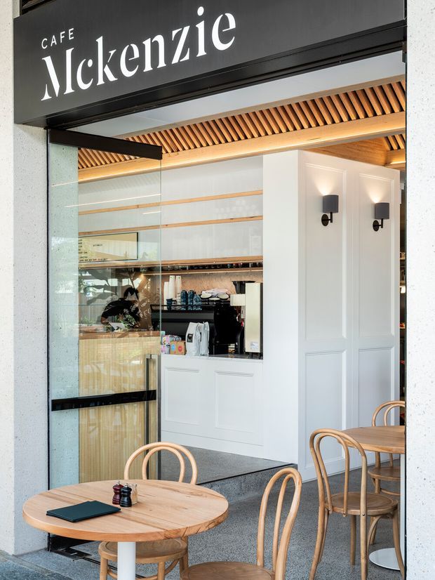 A refined cafe entrance and interior viewed from the exterior. A black sign reading “Cafe McKenzie” sits above full-height glazing and a dark-framed entry. Inside are a white service counter, espresso machine, light timber shelving and panelling, decorative wall sconces, and a slatted timber ceiling with warm integrated lighting. Round light-wood tables and bentwood chairs occupy the foreground, creating a bright, contemporary hospitality setting.