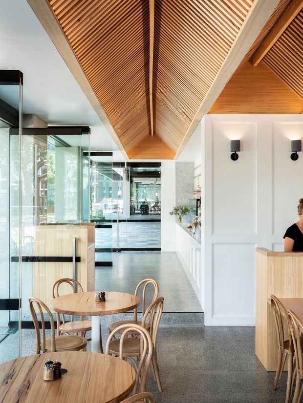 A bright contemporary café or hospitality interior with round timber dining tables and bentwood chairs in the foreground. The space features a dramatic pitched timber ceiling lined with closely spaced slats and exposed ridge structure, polished concrete or terrazzo flooring, extensive glass partitions, white decorative wall panelling, and a light timber service counter. A long open view extends through the dining area toward glazed doors and an outdoor setting.