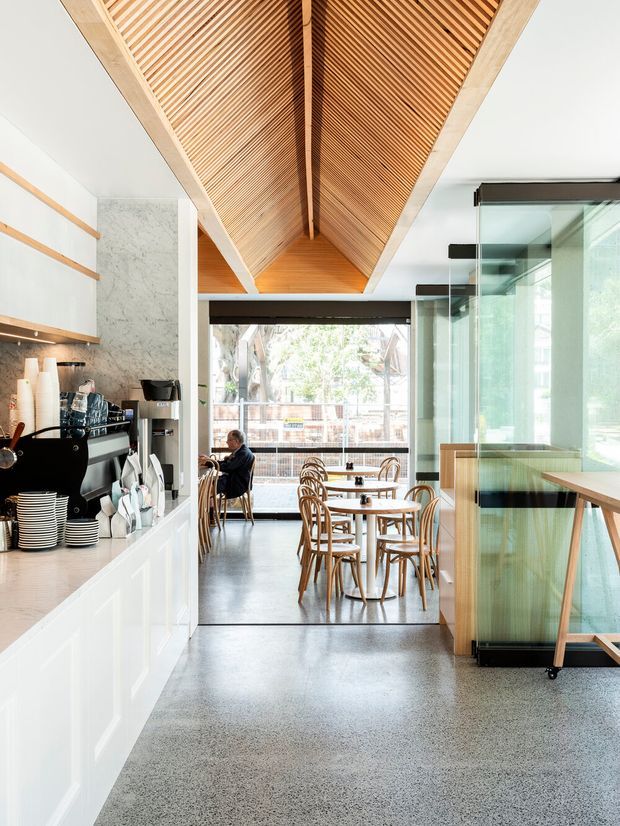 A bright contemporary café interior viewed from the service counter toward a dining area. The space features a long white counter with coffee equipment, a grey marble backsplash, polished speckled flooring, timber café tables and bentwood chairs, extensive glazing, and a dramatic pitched ceiling lined with closely spaced timber slats and a central exposed beam.