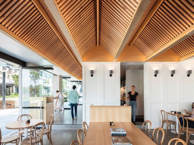 A bright contemporary hospitality interior, likely a café or restaurant, with timber dining tables and bentwood chairs, expansive floor-to-ceiling glazing, white decorative wall panelling, and a striking pitched ceiling lined with exposed timber battens and beams. The space has a warm timber-and-white palette and a strong connection to the outdoors through the glazed frontage.