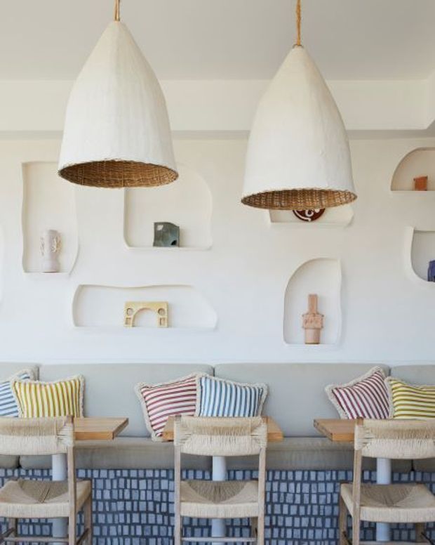 Bright, playful coastal-style lounge or hospitality seating area with a long built-in upholstered bench, woven timber-framed chairs, striped cushions, and a blue patterned tile or textile-like lower wall. The white sculptural wall with recessed display niches is complemented by two large woven pendant lights and small decorative objects.