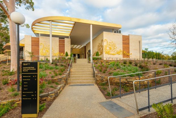 Exterior view of a contemporary public building entrance with a broad curved canopy, pale rendered walls, warm timber cladding, decorative golden wall artwork, landscaped planting beds, stairs and accessible ramping. A wayfinding sign identifies the Central Park Room and Function Room, indicating a civic or commercial venue.
