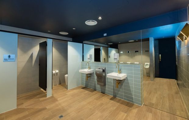Contemporary commercial public bathroom interior with multiple toilet cubicles, wall-mounted basins, large mirrors, pale blue tiled half-height partitions, beige wall tiles, warm wood-look flooring, dark ceiling, and blue accent walls. The space is clean, spacious, and evenly lit.