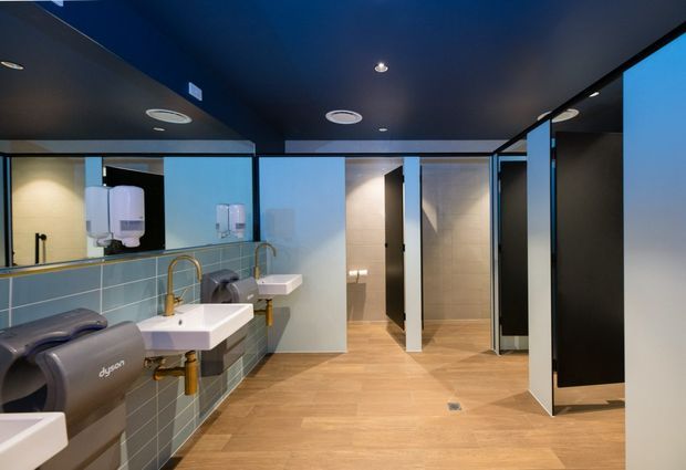 Contemporary commercial bathroom interior with multiple white wall-mounted basins, exposed plumbing, toilet cubicles, hand dryers, large mirrors, and pale timber-look flooring. The space has a dark navy ceiling, black-framed glazing and partitions, grey-green tiled wall surfaces, and warm recessed lighting.