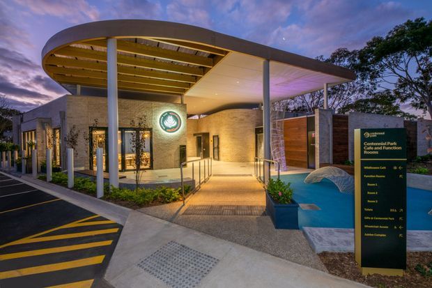 Dusk exterior view of a contemporary public recreation and community facility. The building has a curved metal canopy with exposed timber-toned soffit, slender white columns, textured masonry walls, glazed entry doors, landscaped planting, accessible ramps and handrails, and a blue water feature beside a freestanding directional sign. The image appears to show the Central Park Conference and Function Rooms entrance.